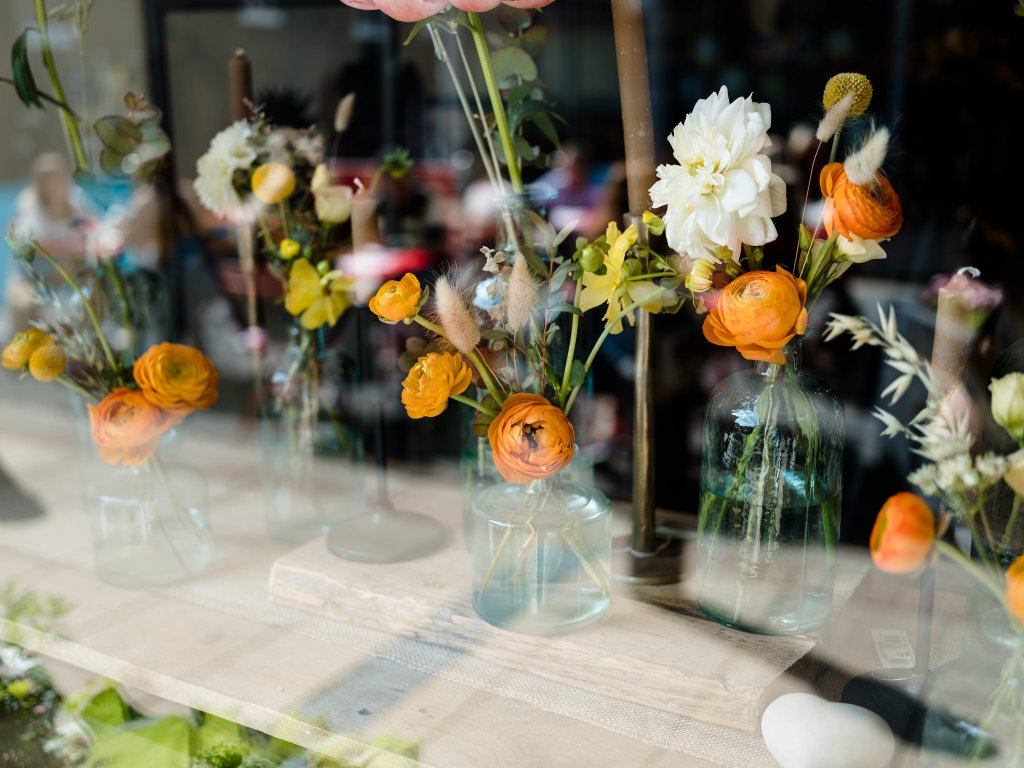small bouquets of orange and white flowers in glass jars seen through a shop window.